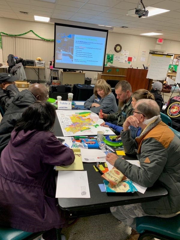 Seven people sit at a table and look over a colorful map. The table is filled with papers, writing supplies, and is located in front of a projector screen.