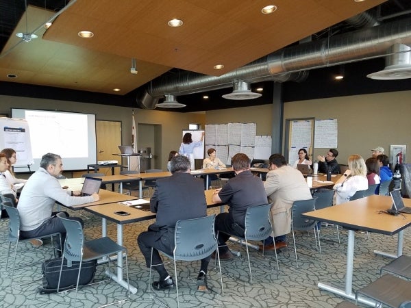 14 people are seated in a round-table focus group discussion. A notetaker writes on a large note pad and additional large pieces of paper are taped onto the walls.