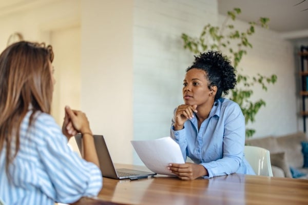Two people sit across from each other for an interview. One person holds a piece of paper and has a laptop in front of them.