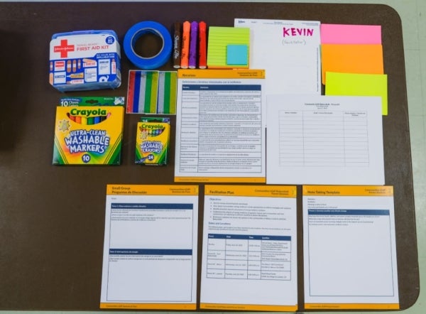 An overhead shot of meeting supplies, including a first aid kit, writing supplies, meeting packets, and tape. These materials are laid out on a table.