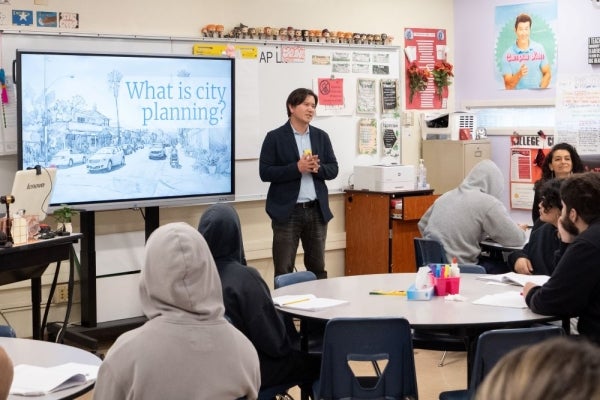 A city planner stands in front of a classroom of youth and next to a presentation that reads "What is city planning?".