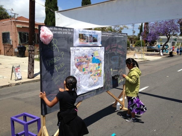 Two children write on a chalk board displaying ideas related to Southeastern San Diego community planning area. The board is outdoors, on a street.