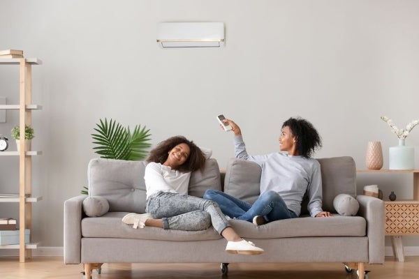 Two people sit on a couch and one person turns on the air conditioning unit.