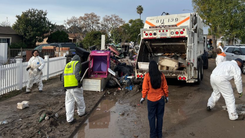 five people working to clear out trash in a trash truck on the street. 