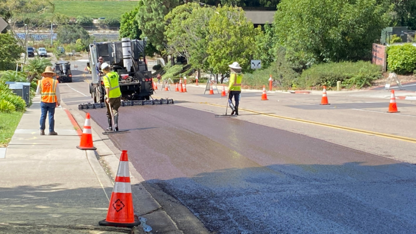three workers slurry sealing a section of a street&#44; surrounded by trees