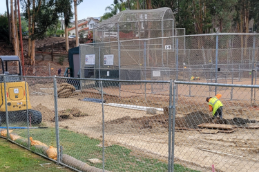 construction worker digging dirt surrounded by a chain fence and baseball field