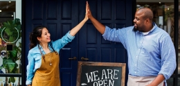 Two small business owners high-five each other in front of a "We are Open" sign.